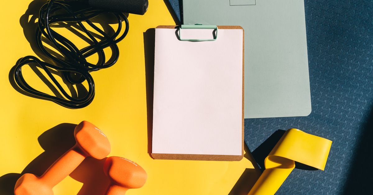 Flat lay of fitness equipment including dumbbells, jump rope, and clipboard on a yellow background.